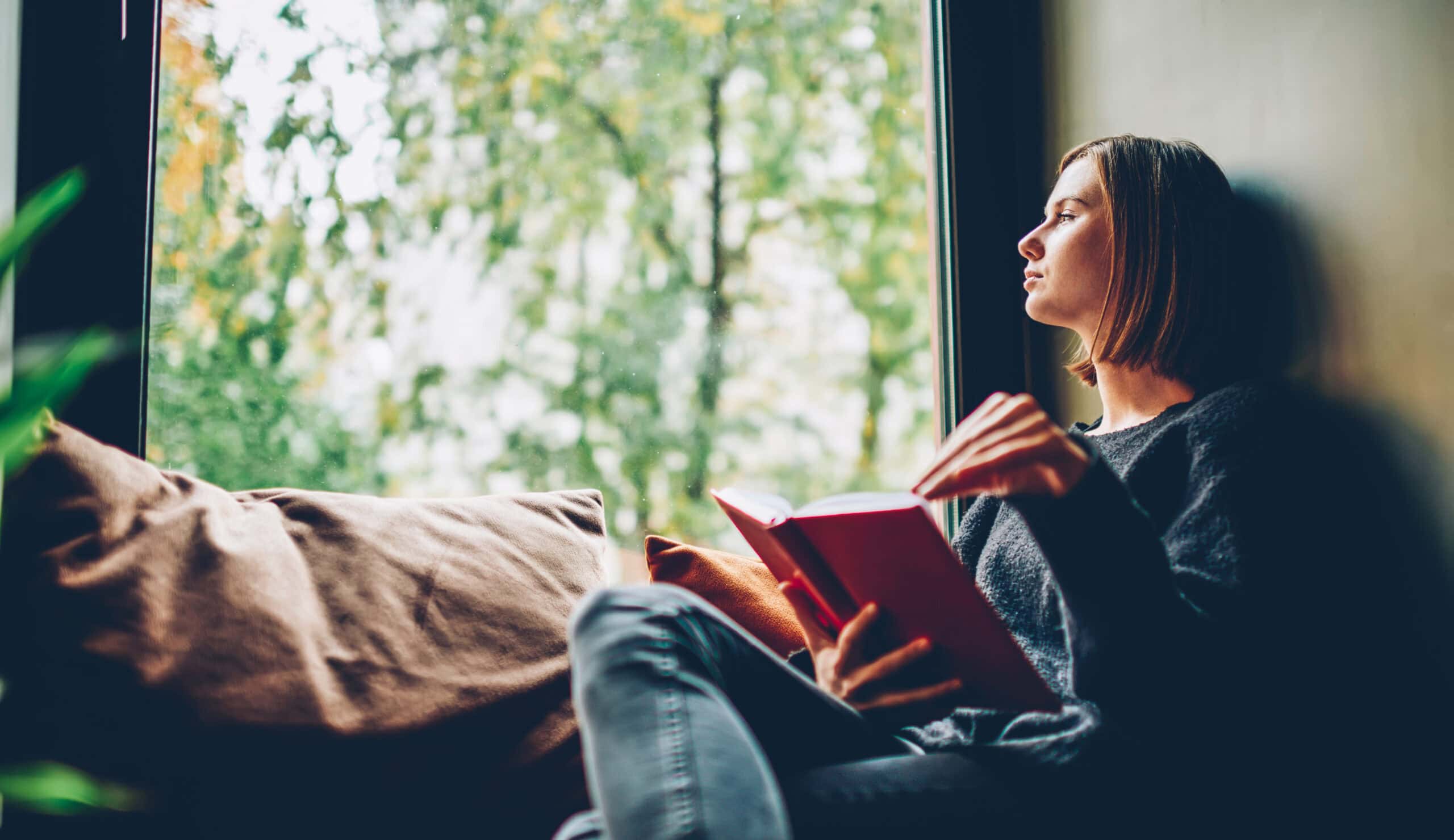 Thoughtful young woman looking out of window pondering on idea while reading a book.